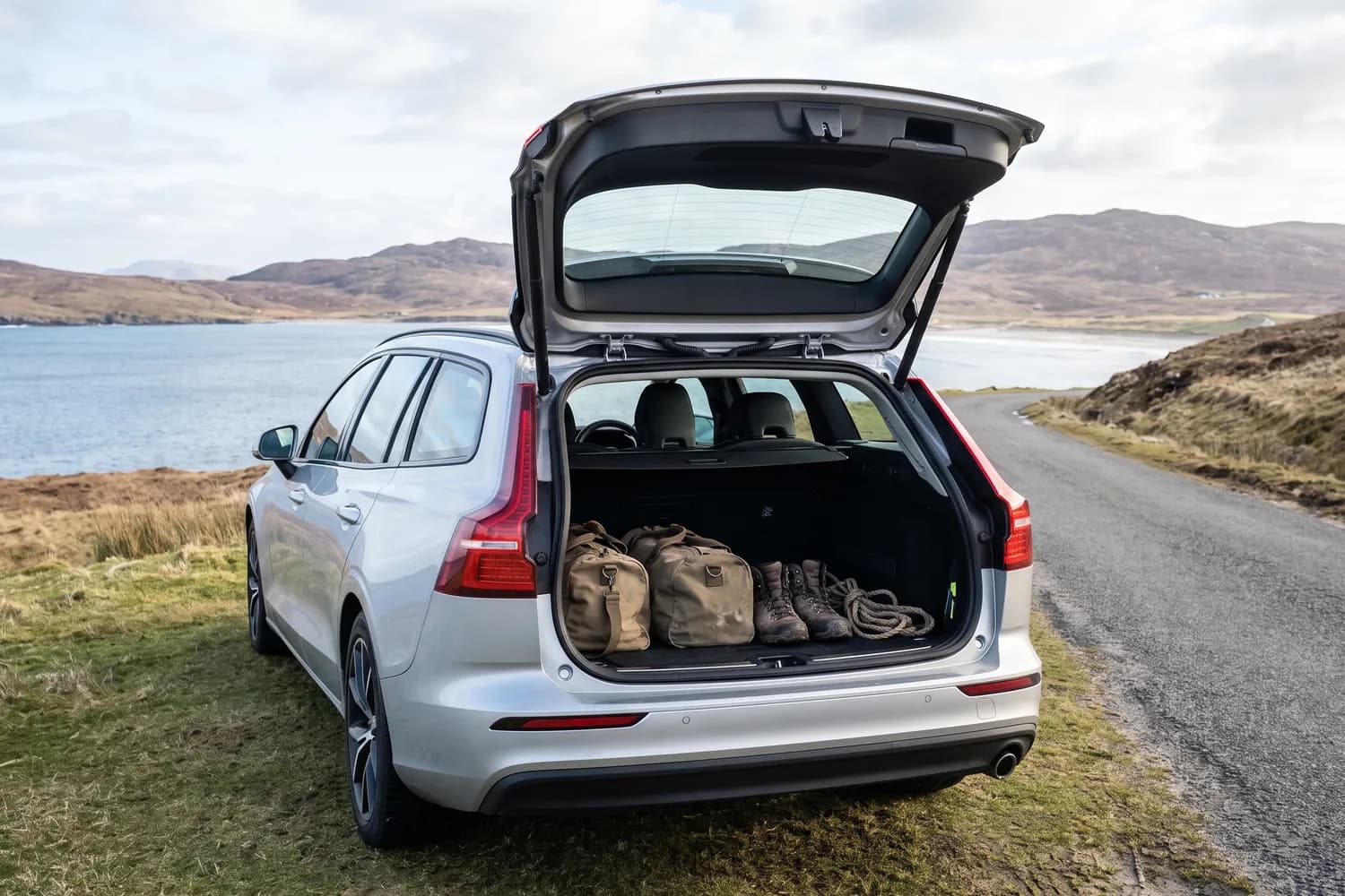 Soft holdalls and walking boots in the open boot of an estate car parked at a roadside above a bay